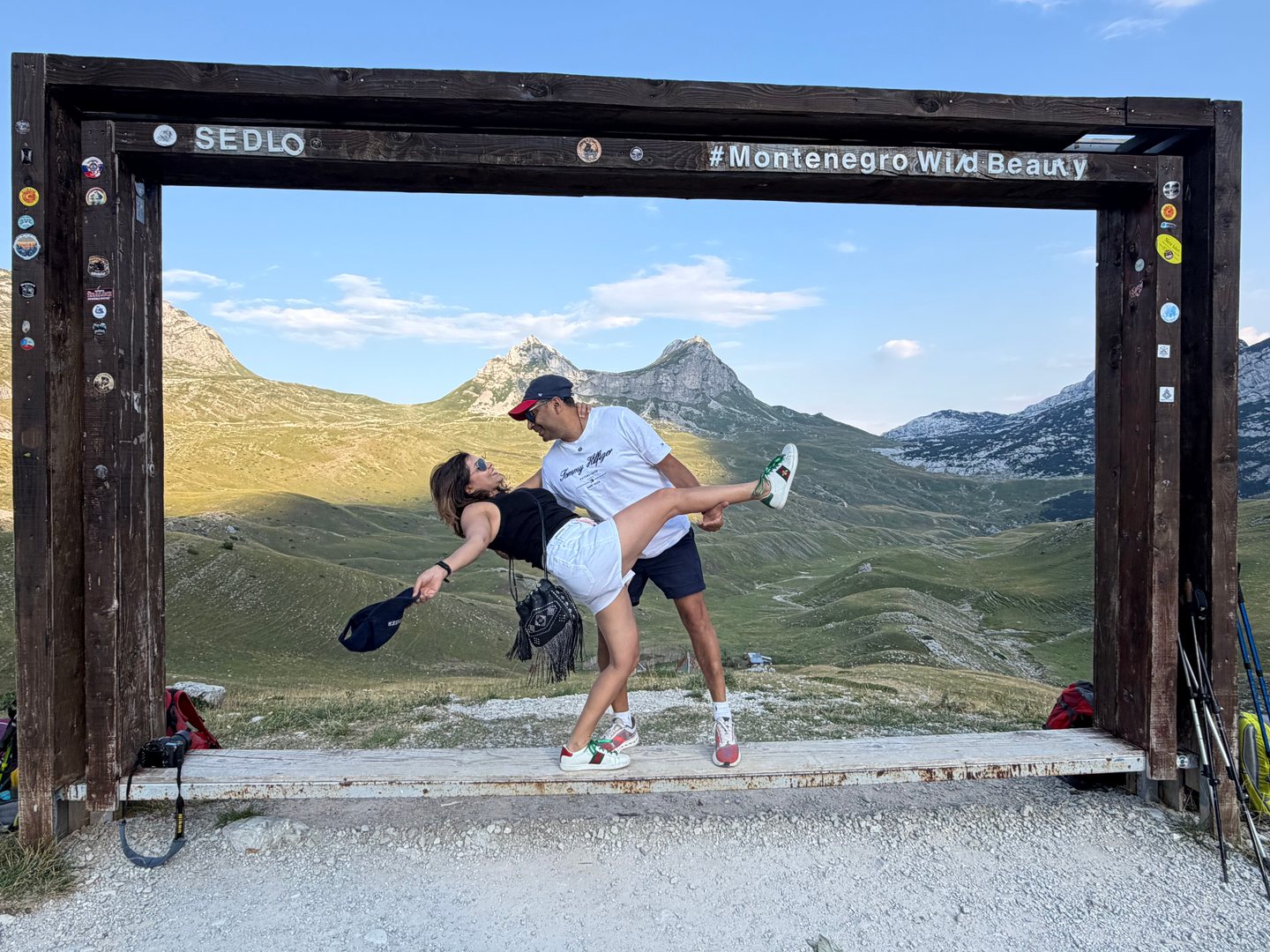 Two adults posing inside a wooden picture frame with rugged green mountains behind them.